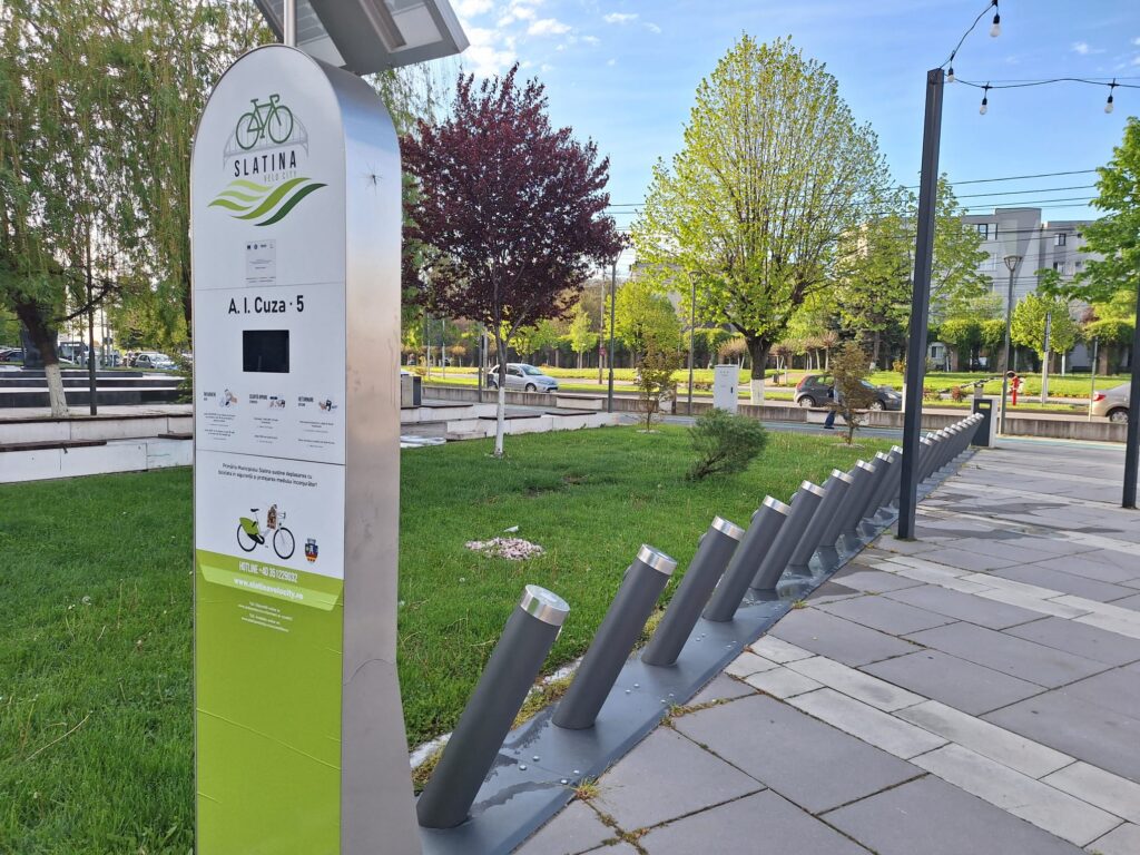 Tall metallic information sign in a park with a bicycle logo and 'SLATINA VELO CITY' text, surrounded by trees and greenery.