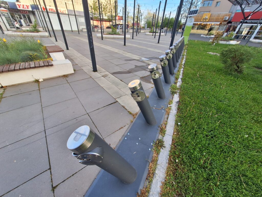 Row of dark vertical posts with silver caps along a paved city plaza, with plants and shops in the background.