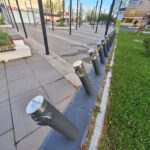 Row of dark vertical posts with silver caps along a paved city plaza, with plants and shops in the background.