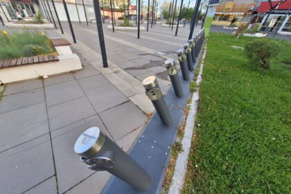 Row of dark vertical posts with silver caps along a paved city plaza, with plants and shops in the background.