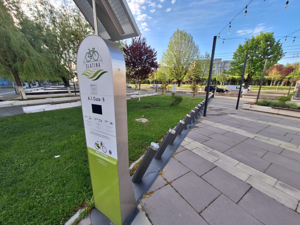 Informational sign about Slatina bike city with bicycle icon and green waves, installed in a grassy park area.