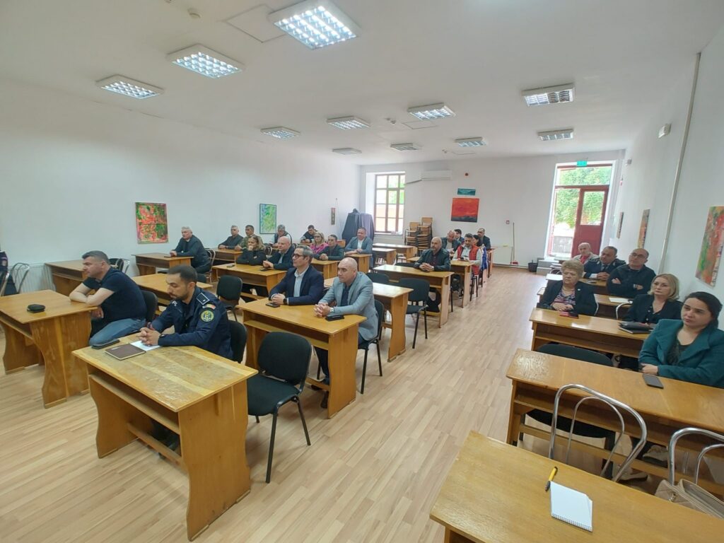 Adults seated at wooden desks in a bright classroom during a meeting or lecture, listening attentively.