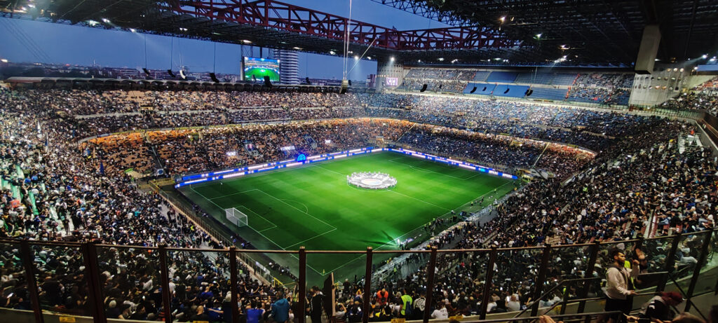 Panoramic view of a packed soccer stadium at night, bright green pitch and fans in the stands.