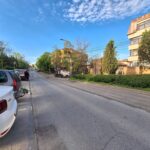 Street view with parked cars along the curb, a beige multi-story building on the right, and a clear blue sky above.
