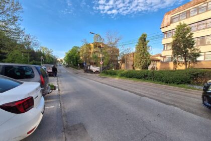 Street view with parked cars along the curb, a beige multi-story building on the right, and a clear blue sky above.