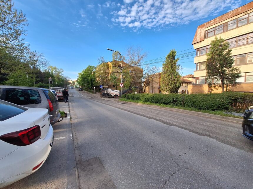 Street view with parked cars along the curb, a beige multi-story building on the right, and a clear blue sky above.