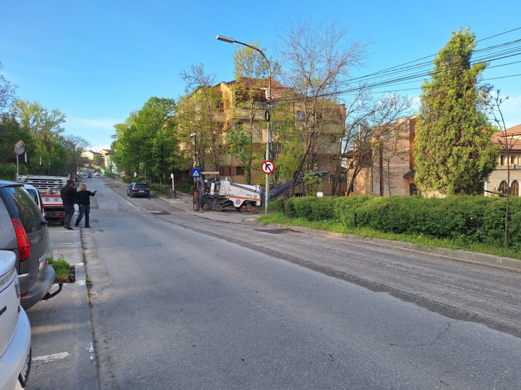 Two men talk on a residential street beside a small construction machine and parked cars under a blue sky with trees nearby.