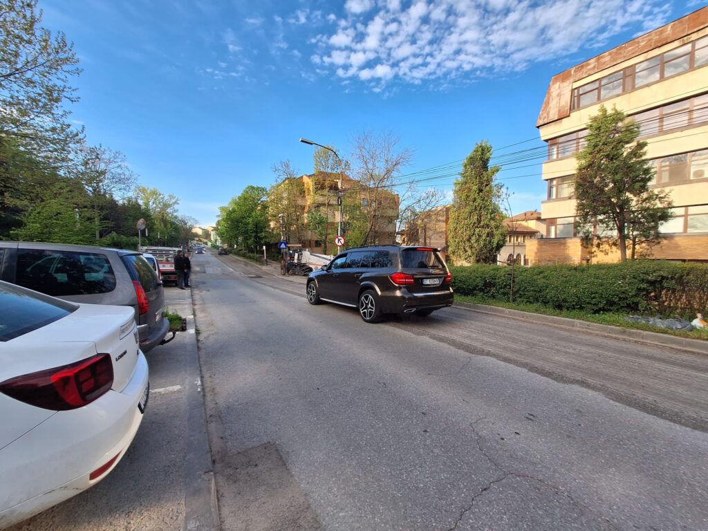 Street scene with parked cars along both sides, a beige multi-story building on the right, and green trees under a blue sky.