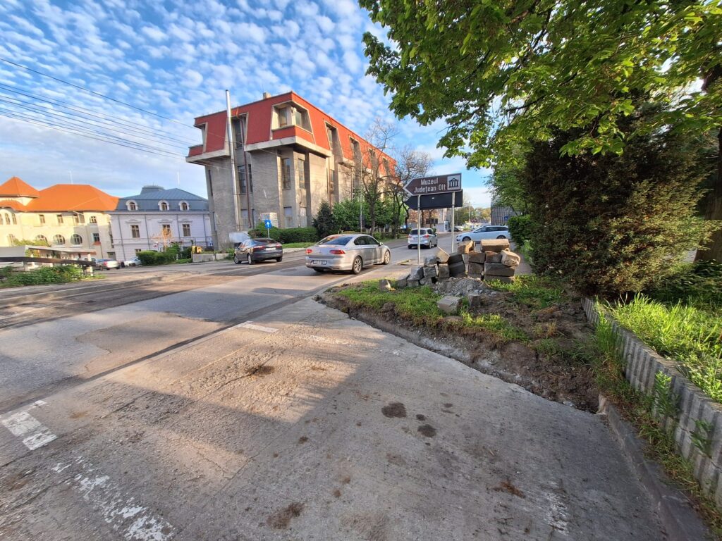 Street scene with a modern red-brick building, parked cars, and a directional sign to Muzeul Judetean Olt; trees frame the right side under a blue sky.