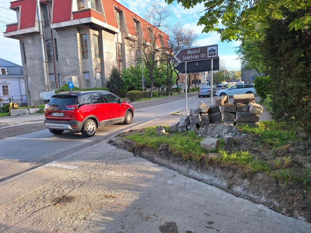 Red SUV on a street with a large pile of concrete blocks and construction debris on the sidewalk nearby.