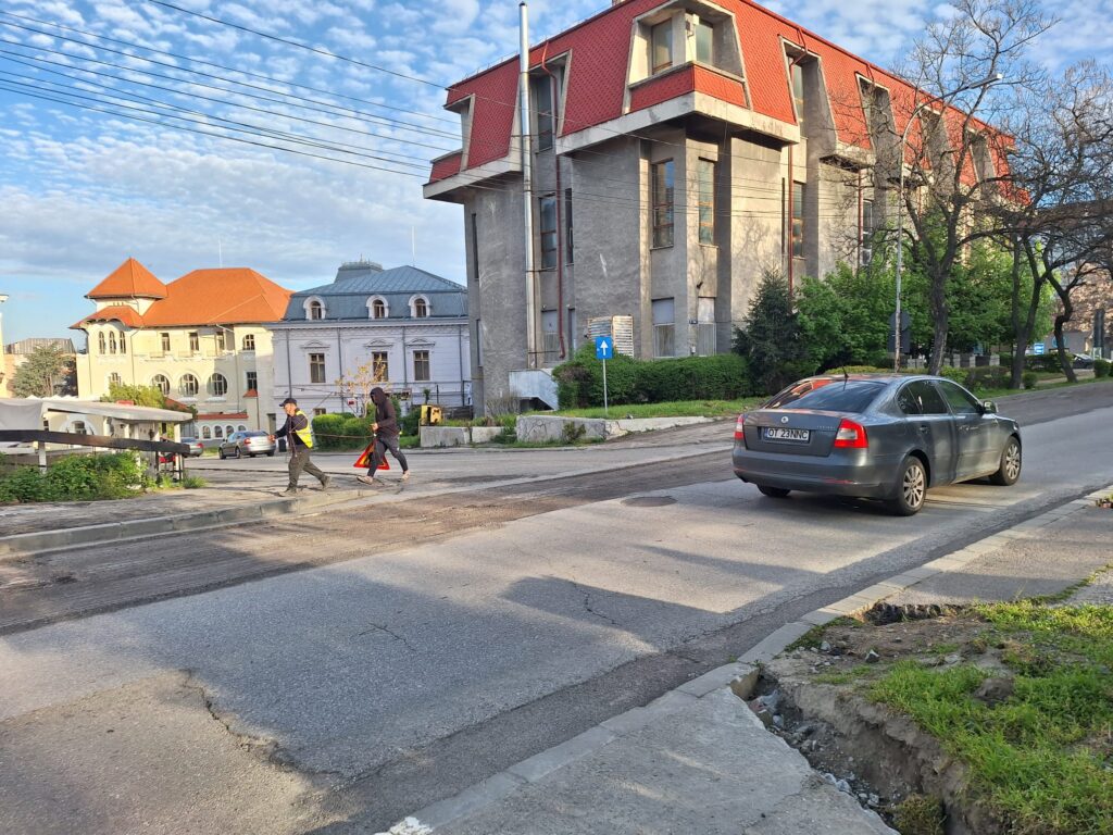Street scene with a grey concrete building under construction, red roof, and two workers in reflective vests near a traffic cone.