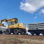Yellow excavator on railroad tracks lifting a stack of concrete railway sleepers beside a flatbed car loaded with sleepers under a blue sky.