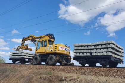 Yellow excavator on railroad tracks lifting a stack of concrete railway sleepers beside a flatbed car loaded with sleepers under a blue sky.