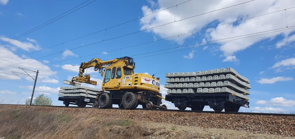 Yellow excavator on railroad tracks lifting a stack of concrete railway sleepers beside a flatbed car loaded with sleepers under a blue sky.