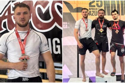 Male athlete posing with a red medal in a white shirt, standing in front of an event banner indoors.