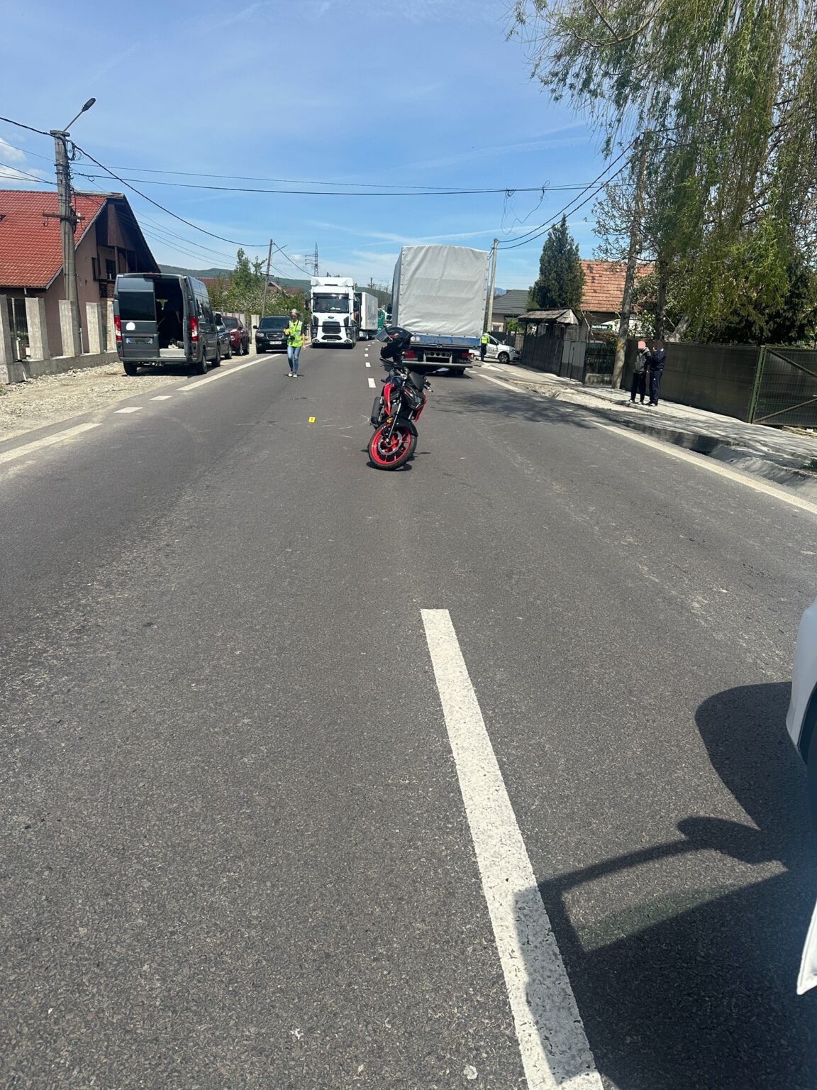 Motorcycle sits in the middle of a residential street with parked vans and trucks along the sides; a person in a high‑visibility vest stands nearby.