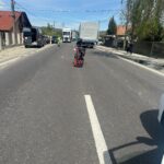 Motorcycle sits in the middle of a residential street with parked vans and trucks along the sides; a person in a high‑visibility vest stands nearby.