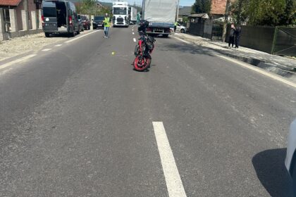 Motorcycle sits in the middle of a residential street with parked vans and trucks along the sides; a person in a high‑visibility vest stands nearby.
