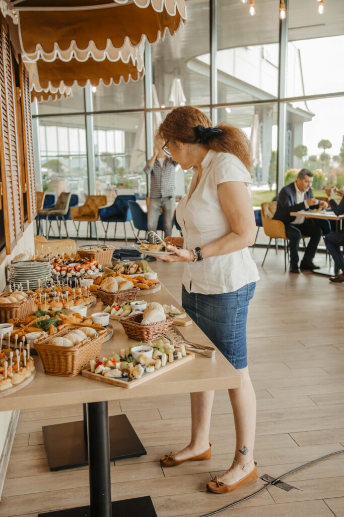 Woman in a white blouse serves herself at a buffet, selecting finger foods from a table of pastries and skewers in a bright cafe-style space.