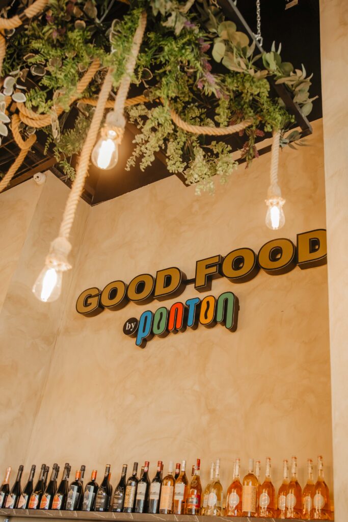 Wall-mounted bar area with hanging rope lights and lush greenery, warm Edison bulbs glowing over bottles of wine and spirits beneath a 'Good Food' sign by pour-toy brand.
