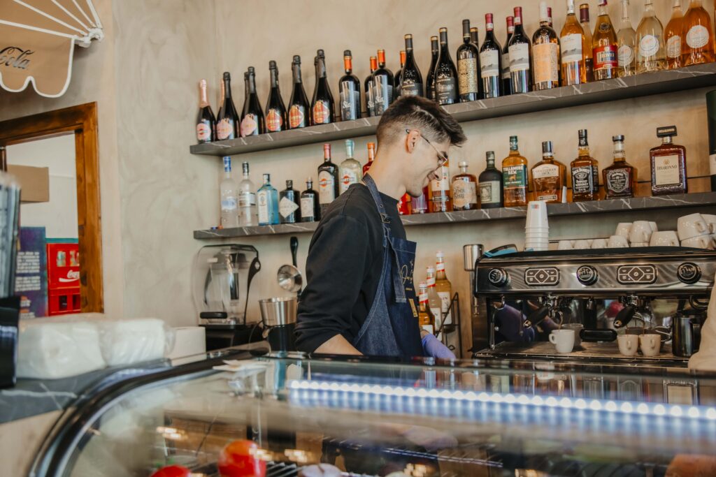 Barista wearing apron and glasses prepares coffee behind a counter with bottles on shelves above him and cups nearby.]','Barista focused on an espresso machine in a cafe, with a wall of bottles on shelves behind.