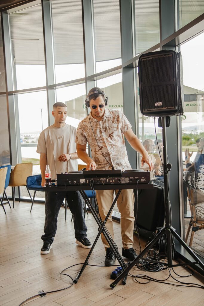 DJ setup indoors with two men at a mixer, headphones on, large speaker nearby and many cables on the floor