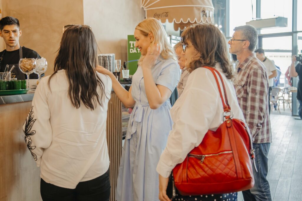Group of people waiting at a cafe counter, some in white coats, with a barista behind the counter and a red quilted handbag in the foreground.