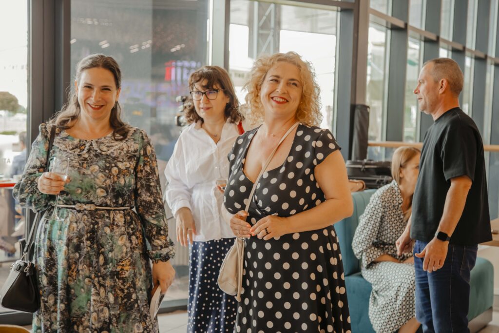 Smiling group of adults at a social event indoors, holding drinks, with two women in patterned dresses in the foreground.