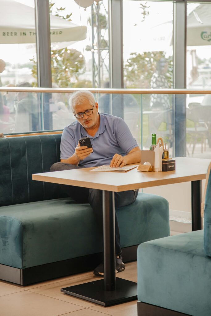 Older man wearing glasses and a blue polo shirt sits in a teal booth at a cafe, looking at his smartphone while a paper menu and condiments sit on the table nearby.