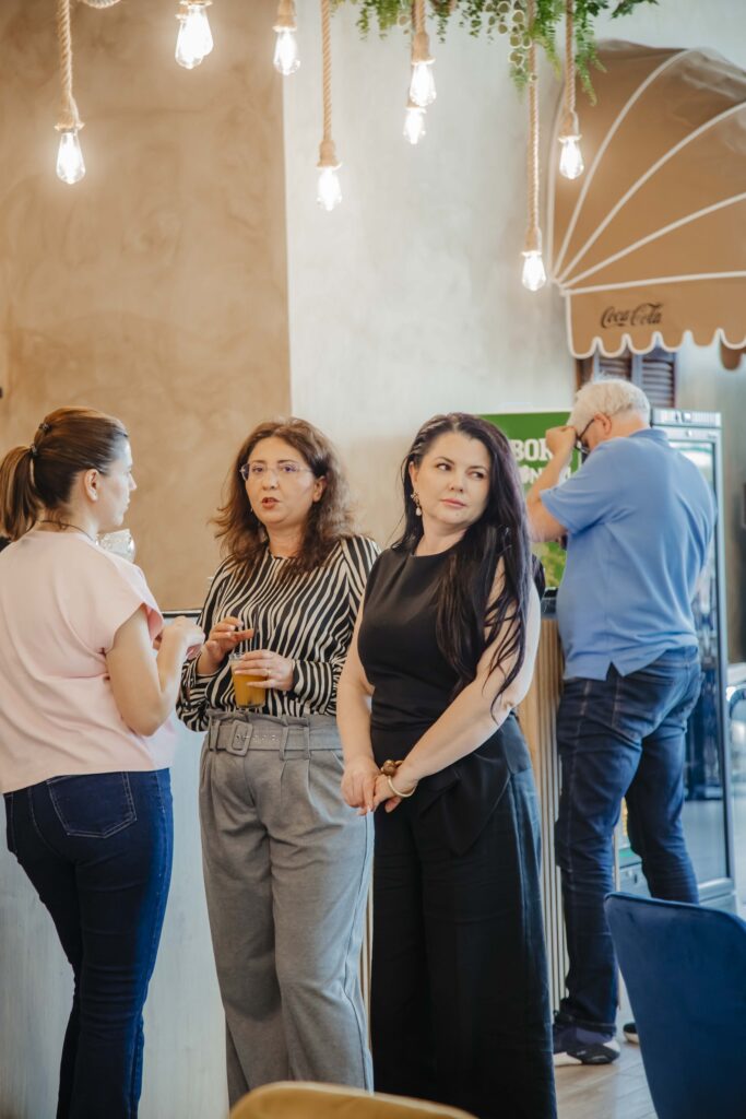 Group of adults chatting at a cafe counter, holding drinks, under string lights and greenery. In the background, a bar area and a Coca-Cola umbrella are visible.