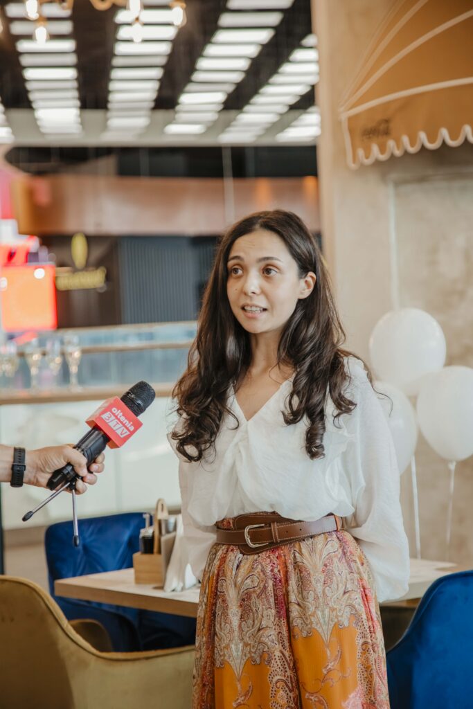 Woman being interviewed indoors, speaking into a handheld microphone held by an unseen reporter, wearing a white blouse and patterned orange skirt.