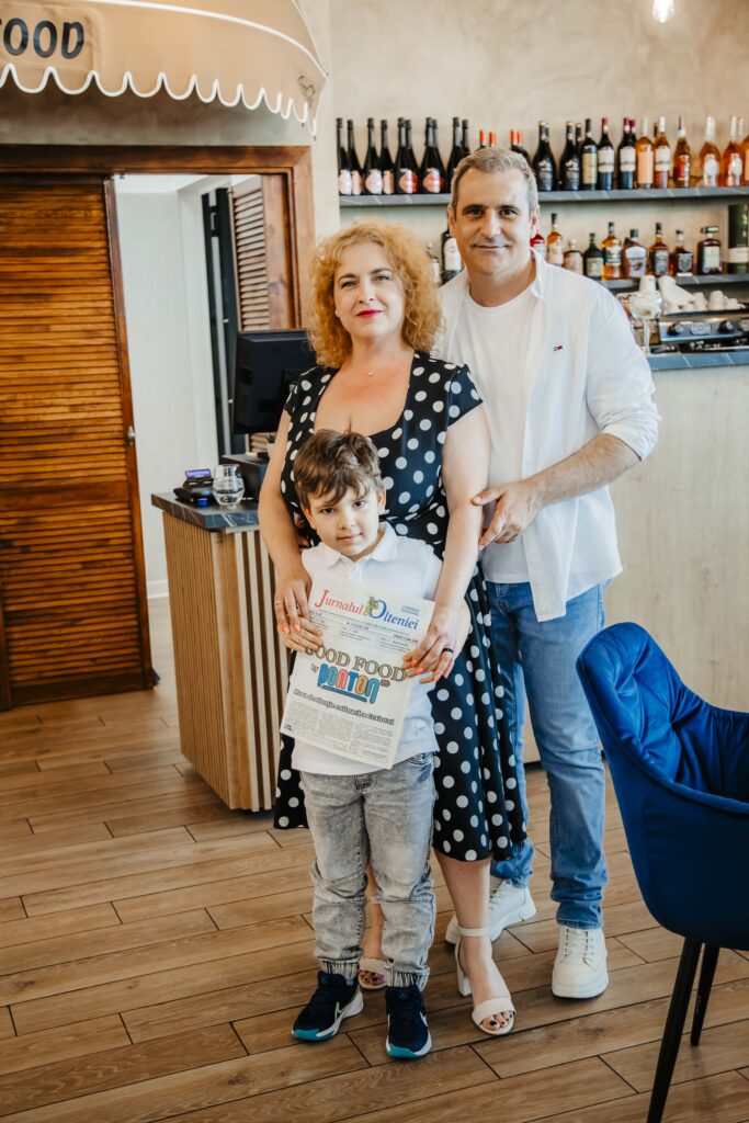 Mother, father, and son pose together in a cafe; the child holds a newspaper while the adults smile behind him.