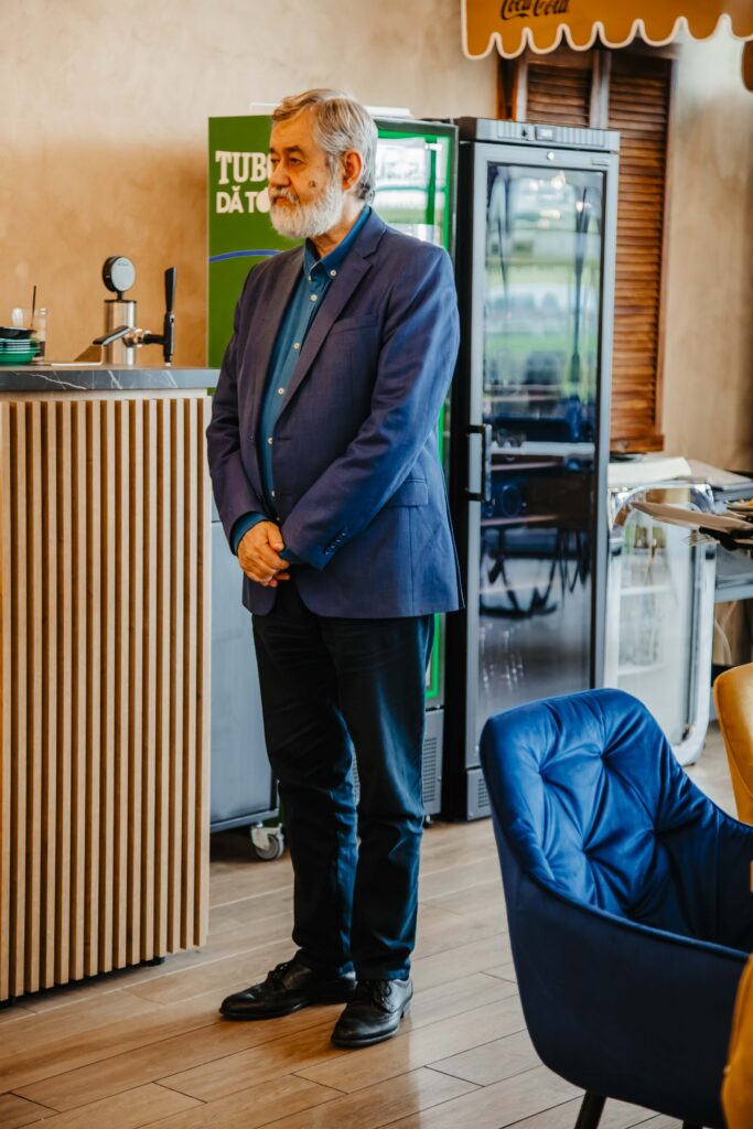 Older man with a gray beard in a blue blazer stands with hands clasped in a cafe, beside a green beverage cooler and a glass-front fridge.