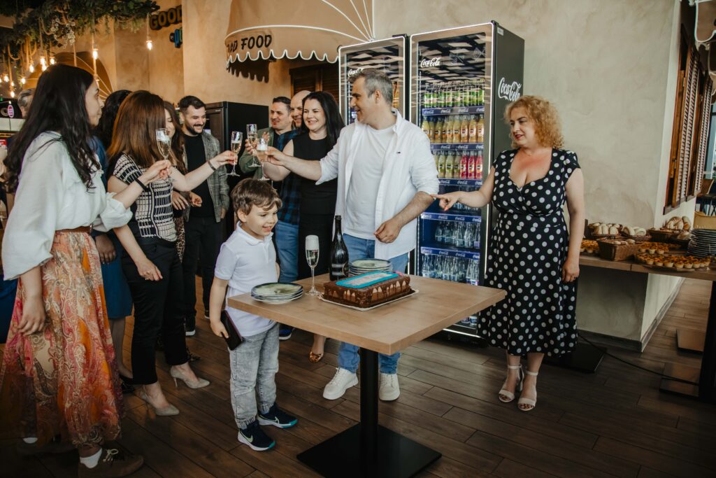 Group of people toasting with champagne glasses around a table with a chocolate cake in a cafe/bakery setting.