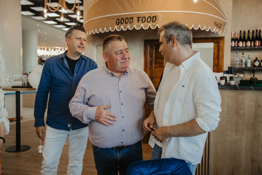 Three men in casual outfits chat and smile in a modern restaurant, with a 'Good Food' canopy overhead behind them.