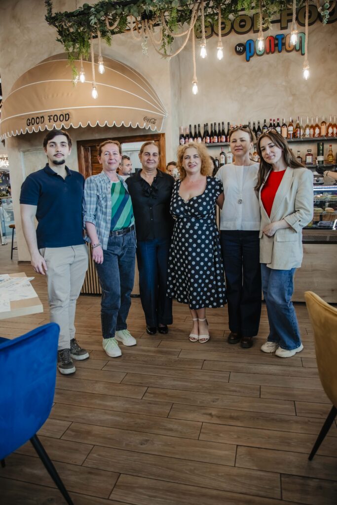 Six adults stand together in a cafe, smiling, behind the bar with bottles and a 'Good Food' sign overhead.