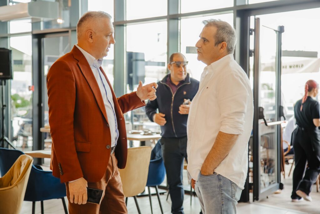 Two men in a casual business chat indoors, one in a rust-colored suit jacket and the other in a light shirt, facing each other.