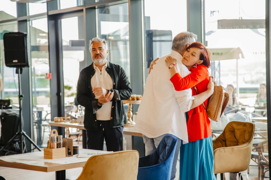 Woman in a red jacket hugs a man in a white shirt at a bright indoor cafe during a social gathering with pastries on the table behind them.