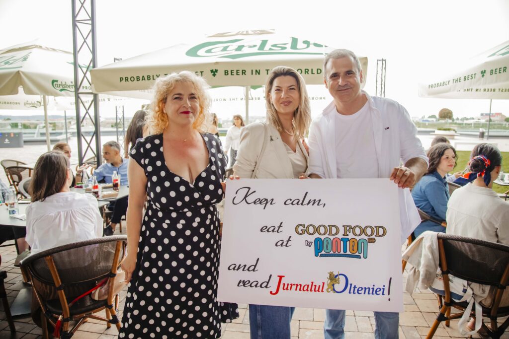 Three smiling adults posing outdoors under Carlsberg umbrellas, holding a large sign promoting good food.