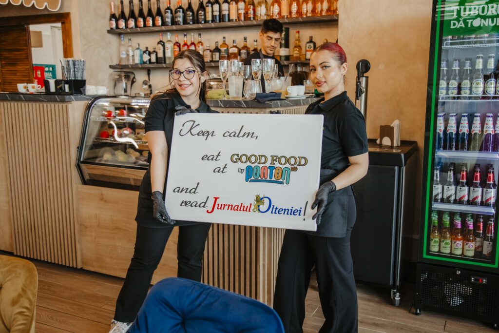 Two cafe/bar staff in black uniforms pose behind a counter, holding a sign that reads “Keep calm, eat GOOD FOOD …” with a bottle display behind them and a fridge to the right.