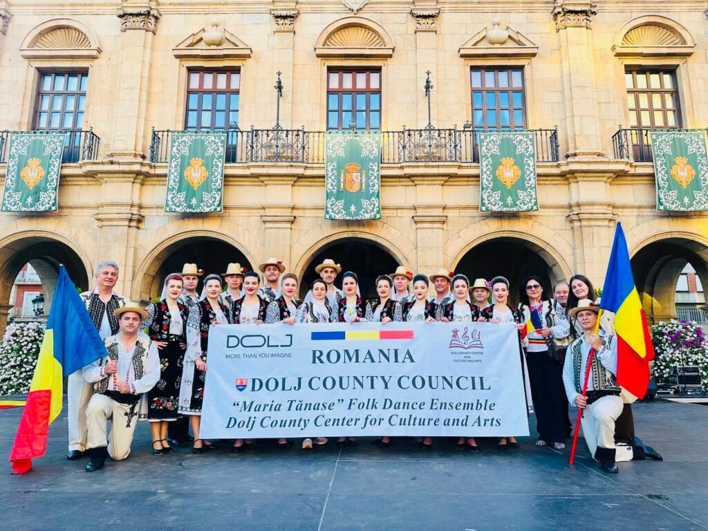 Group of people in traditional Romanian folk costumes pose with a banner for Dolj County Council and Romania flags in front of a historic building with arched entrances and green banners above.