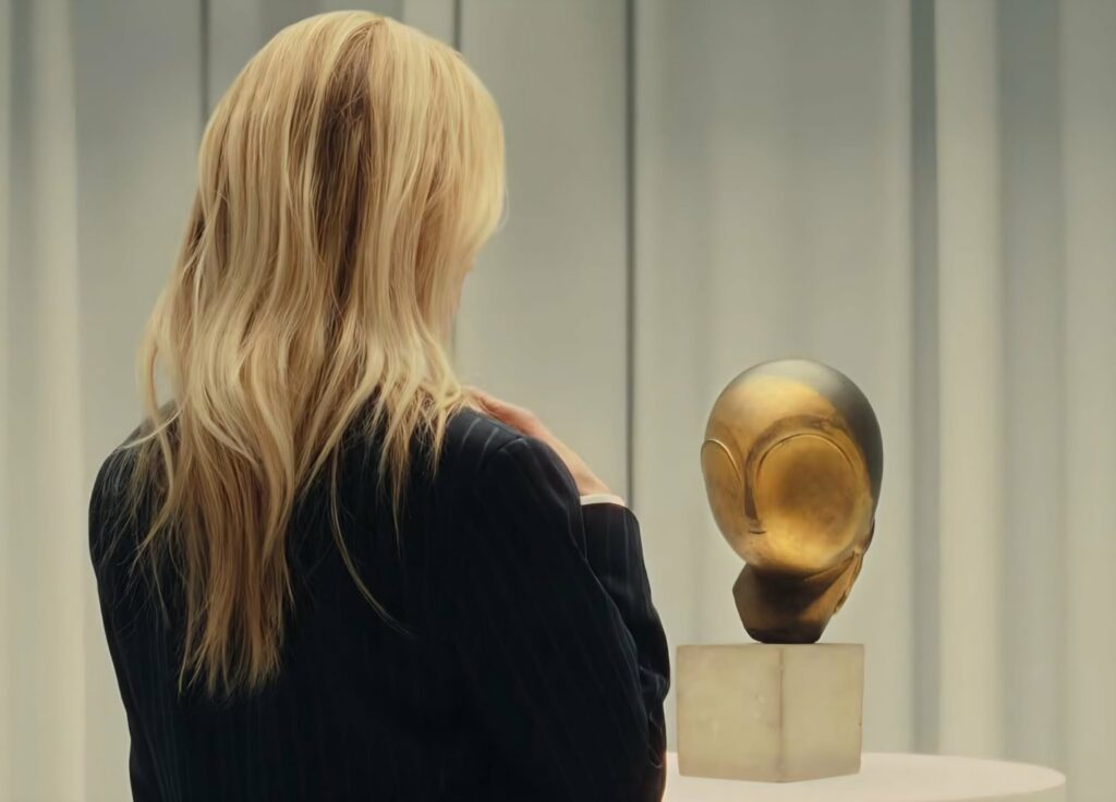 Back view of a blonde woman in a dark jacket looking at a wooden sculpted head on a pedestal in a gallery
