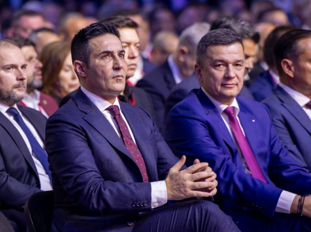 Two men in suits sitting in the front row of a formal event, listening attentively.