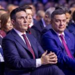 Two men in suits sitting in the front row of a formal event, listening attentively.