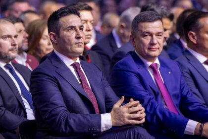 Two men in suits sitting in the front row of a formal event, listening attentively.