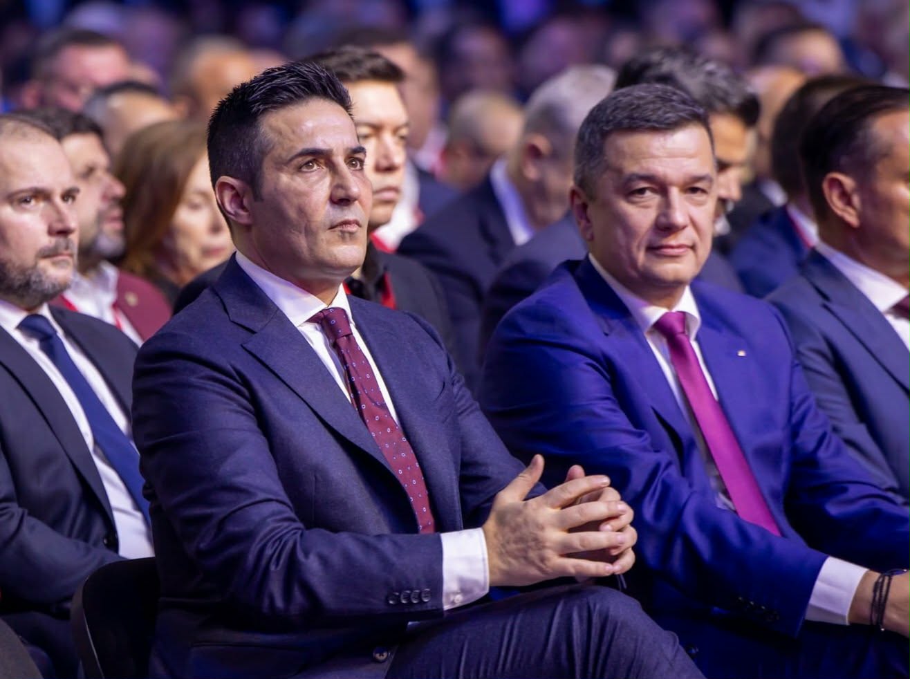 Two men in suits sitting in the front row of a formal event, listening attentively.