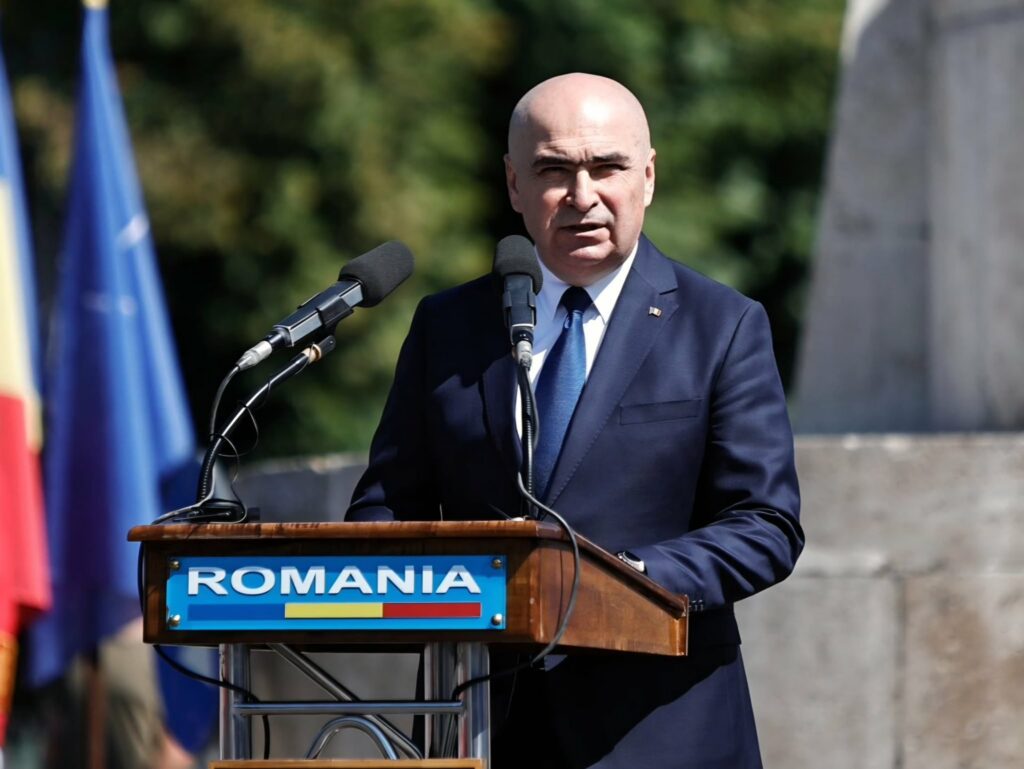 Ilie Bolojan. Foto: Facebook - Jurnalul Olteniei – Știri din Craiova și Oltenia Man in a navy suit speaks at a wooden podium with microphones outdoors, Romania sign in view