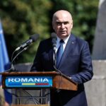 Man in a navy suit and blue tie speaks at a wooden podium labeled ROMANIA, with multiple microphones in front of him.