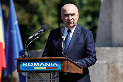 Man in a navy suit speaks at a wooden podium with microphones outdoors, Romania sign in view