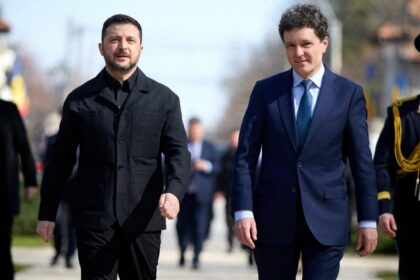 Two men in formal attire walk outside side by side, one in a black jacket and the other in a navy suit, with a blurred background behind them.